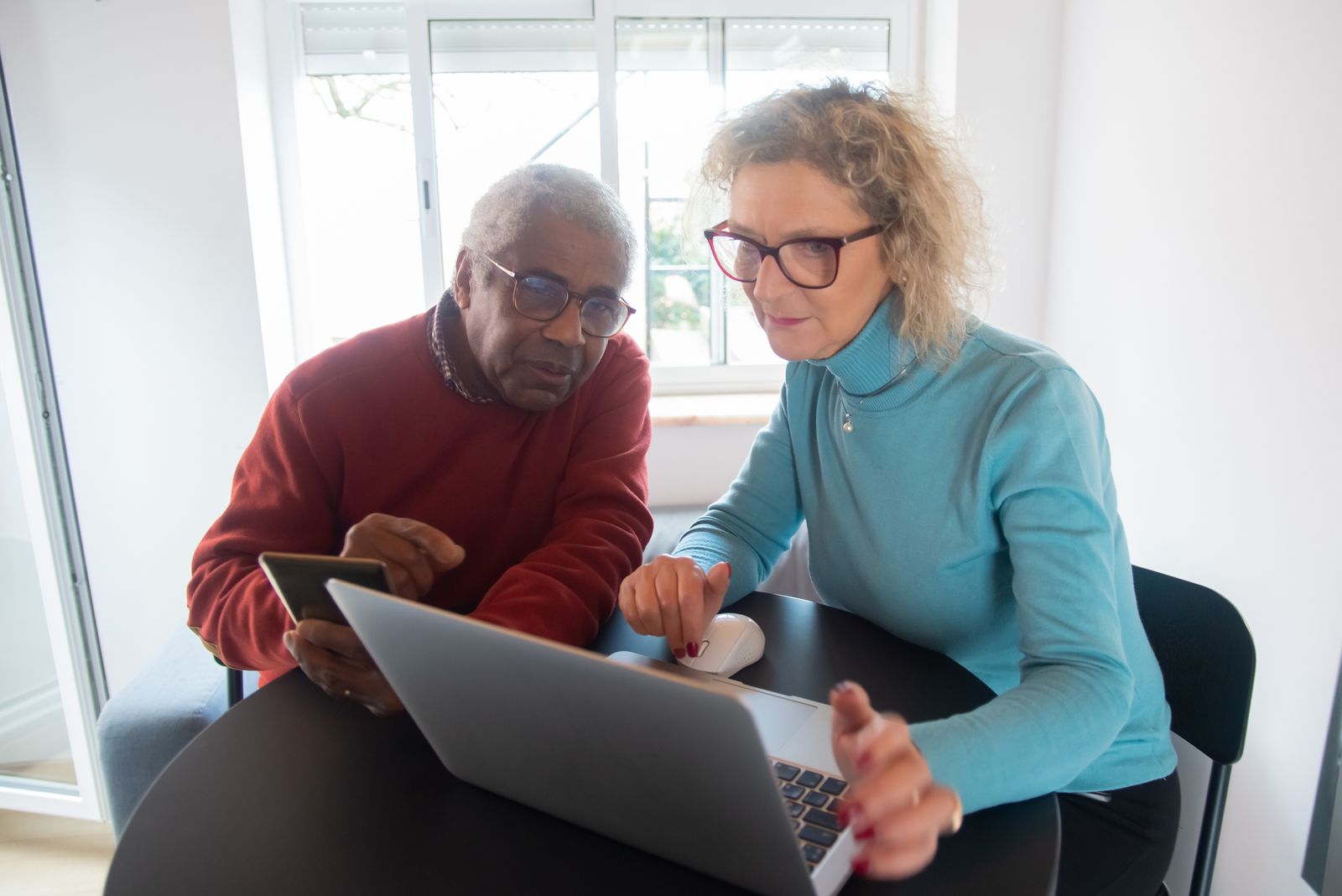 An older couple looks at a laptop together at a table by a bright window